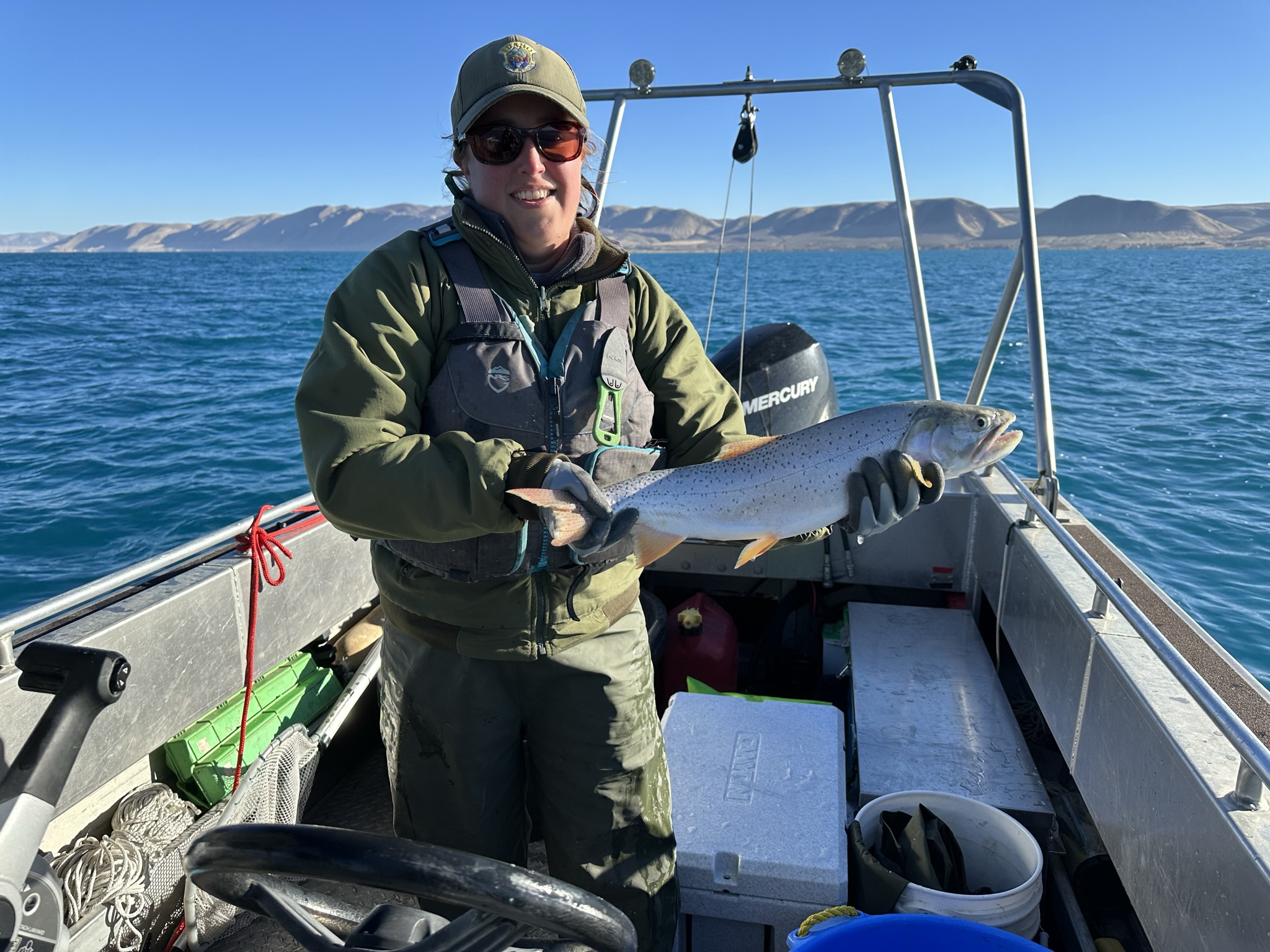 A woman standing in a large jet boat on a very blue lake holding a large silvery cutthroat trout.