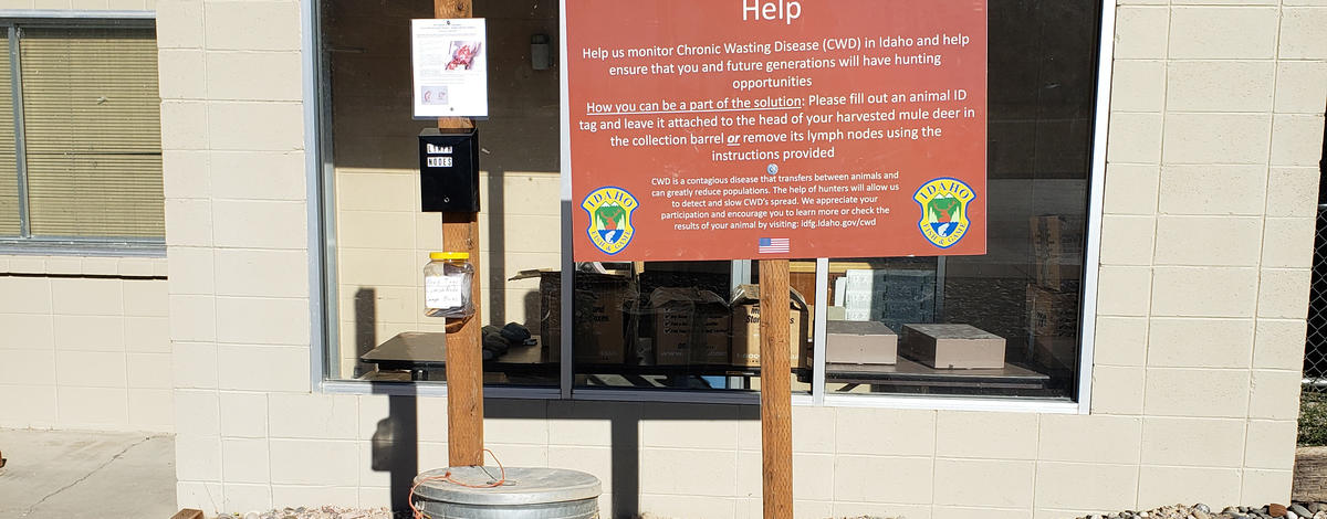 A garbage can next to a large sign with instructions asking mule deer hunters to deposit Chronic Wasting Disease samples in the can.